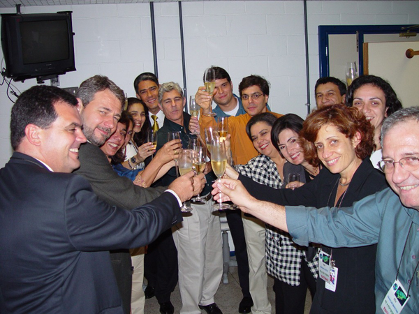 ali kamel e equipe de cobertura, 2002, 3-10 - Ali Kamel (de laranja, ao centro), diretor-executivo de jornalismo, brinda com a equipe o sucesso da cobertura eleitoral de 2002. No 
canto inferior direito, o jornalista Franklin Martins, então diretor da Gl